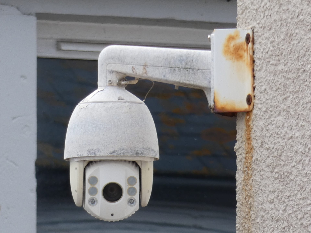 Dusty, white security camera mounted on a rusty white bracket attached to a textured, light beige wall. The camera is a dome-style PTZ (pan-tilt-zoom) model, indicating its ability to move and adjust its view. The overall condition suggests age and exposure to the elements.