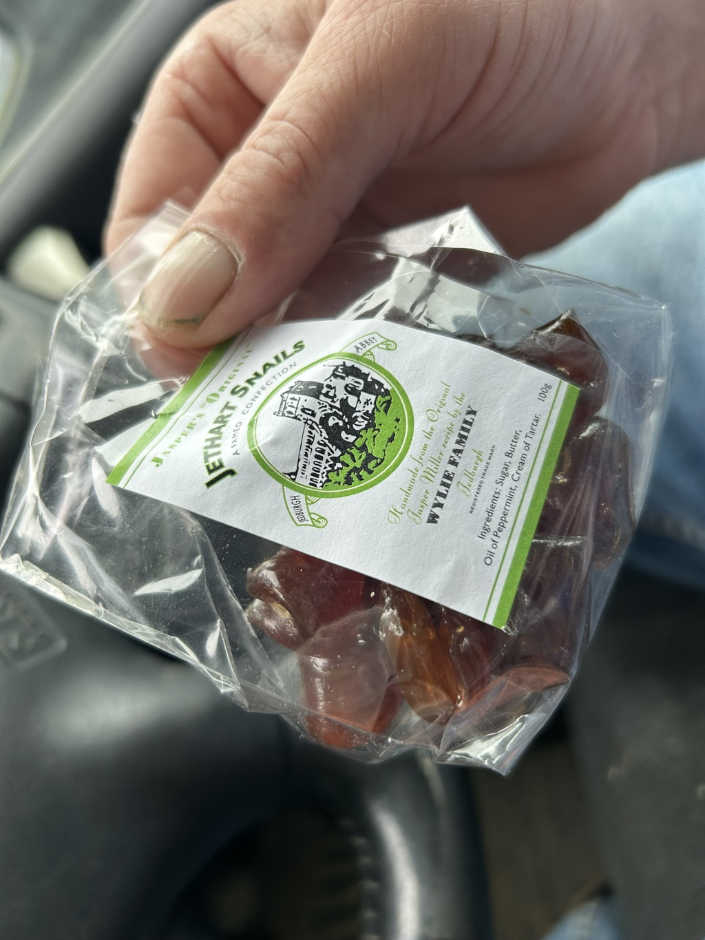 Charlie's hand holding a small, transparent plastic bag containing several pieces of dark brown, irregularly shaped hard candy. The bag has a label indicating that the candy is called Jethart Snails, a famed confection from Jedburgh, Scotland, handmade by the Wylie family using an original Jasper Miller recipe. The label also lists the ingredients. The overall impression is of a simple, perhaps artisan, sweet treat.
