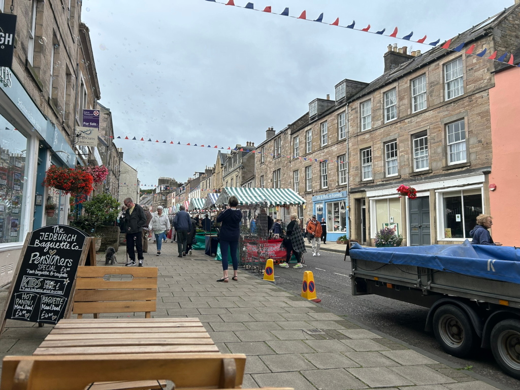 Street scene in a town, possibly in the UK, judging by the architecture and signage. A market is taking place, with several market stalls set up along the street under striped awnings. People are strolling along the pavement, browsing the stalls, and doing some shopping. There's a chalkboard menu for a Jedburgh Baguette Co visible in the foreground. The street is lined with stone buildings, some showing signs of businesses. Festive bunting hangs across the road, indicating a possible local celebration or event. The overall atmosphere is calm and pleasant, suggestive of a community gathering on a fairly ordinary day.
