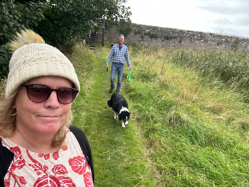 Selfie of Leonie wearing a cream-coloured knit hat and sunglasses. She is standing on a grassy path, with Charlie and a black and white border collie walking away from her in the background. The path leads toward a stone wall. The overall scene suggests a rural or countryside setting; a casual walk in nature.