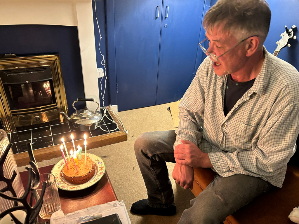 Charlie sitting on a brown leather couch, looking at a small birthday cake with lit candles on a plate in front of him. The setting appears to be a cosy living room with a fireplace, a kettle, and a banjo visible in the background. Charlie is wearing a plaid shirt and jeans. The overall mood suggests a quiet, intimate birthday celebration.