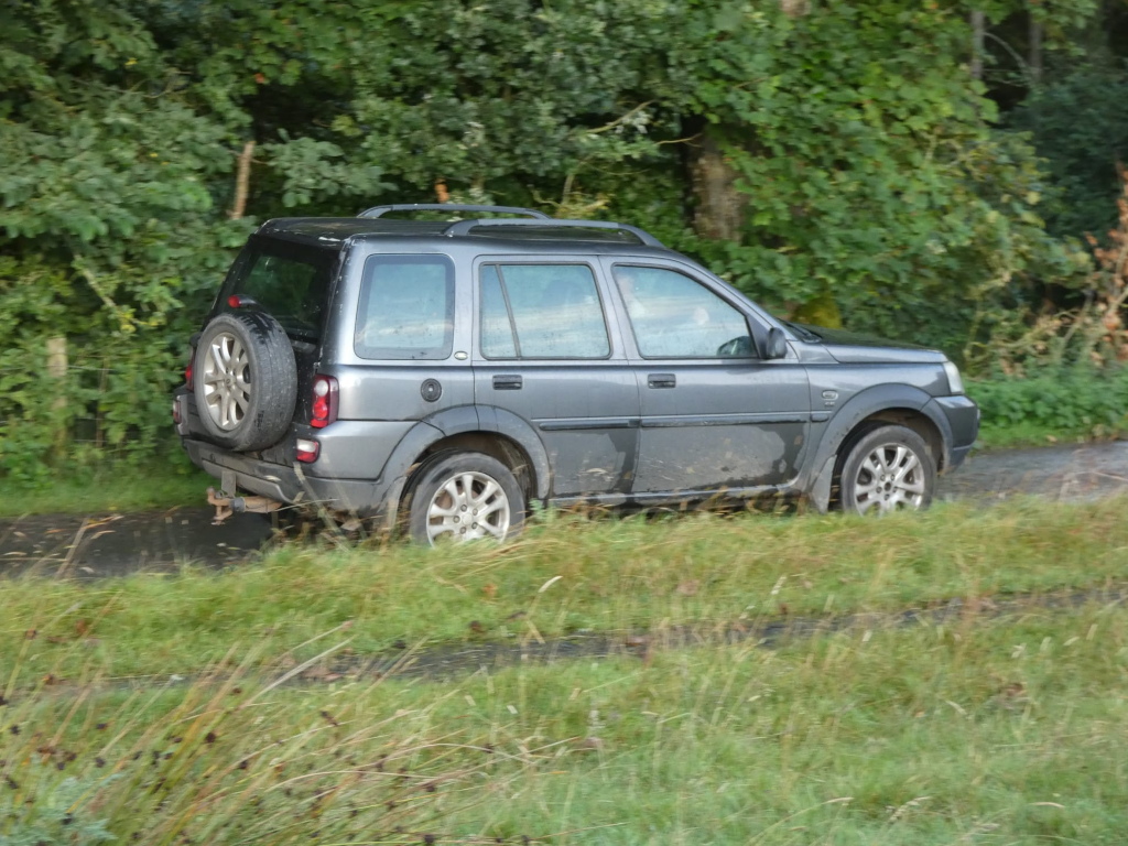 Dark grey Land Rover Freelander driving on a narrow, unpaved road that cuts through a lush green landscape. The vehicle appears to be slightly muddy, suggesting it may have recently travelled on an off-road surface. The background consists of dense, overgrown vegetation, typical of a rural or countryside setting. The overall impression is one of a journey through nature.