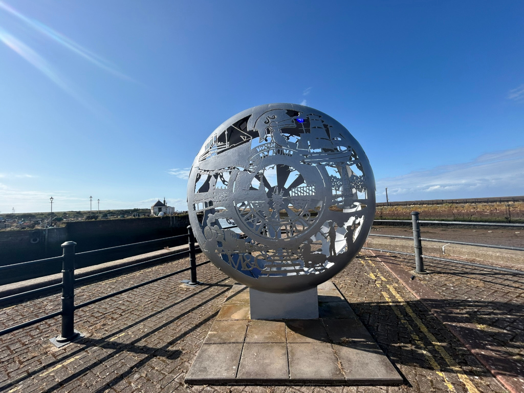 Large, oval-shaped metal sculpture. The sculpture is perforated with cut-out designs depicting various scenes of maritime history, including ships, a wheel, and other nautical imagery. It stands on a stone pedestal in a paved area next to a waterfront. The background includes a waterway, a low wall, and a clear blue sky. The overall impression is one of celebrating a town's seafaring past.