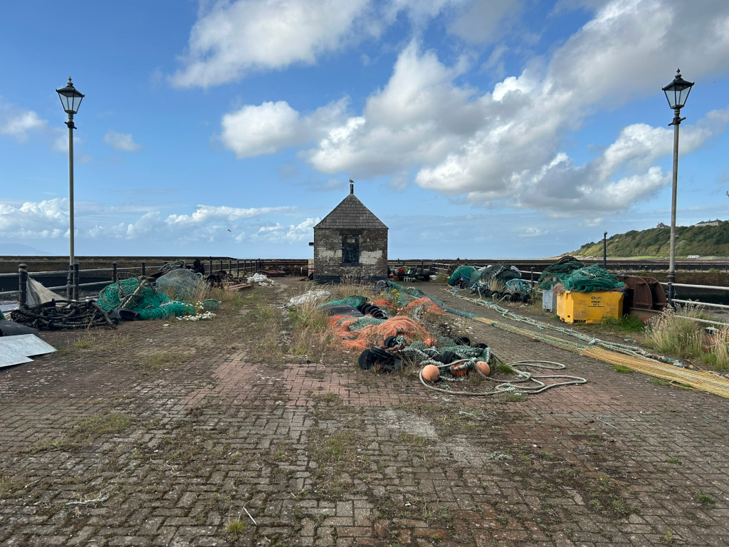 Weathered, brick-paved area, likely a harbour or dock, strewn with discarded fishing nets, ropes, and equipment. A small, aged stone building stands centrally in the background. Two old-fashioned lampposts flank the scene, which is under a mostly sunny, partly cloudy sky. The overall impression is one of quiet abandonment or disuse, hinting at a former bustling fishing activity.