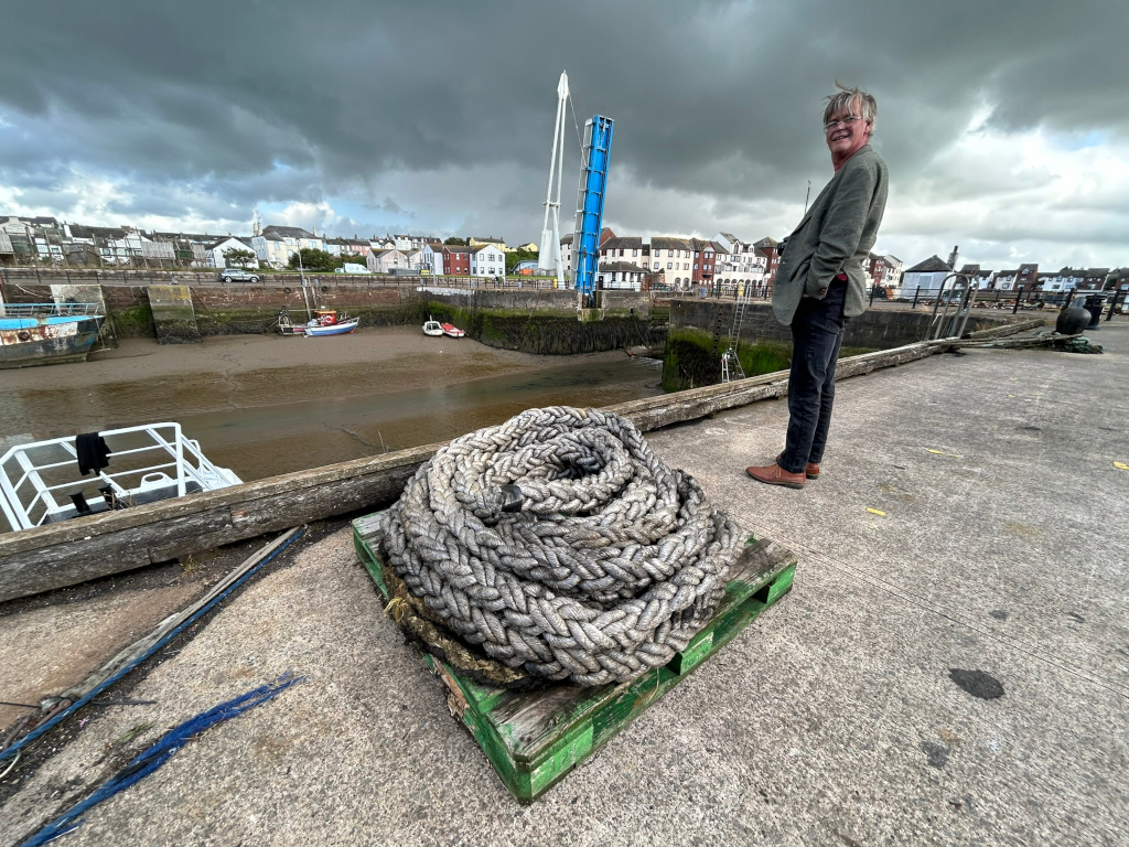Charlie standing on a dock overlooking a harbor. A large coil of thick, weathered rope sits on a pallet in the foreground. In the background, several small boats are moored in the harbour, and a partially cloudy sky looms overhead. The overall scene is one of a quiet, possibly overcast day at a working harbour or marina. Charlie appears to be observing the harbour scene. The buildings in the background suggest a small coastal town or village.