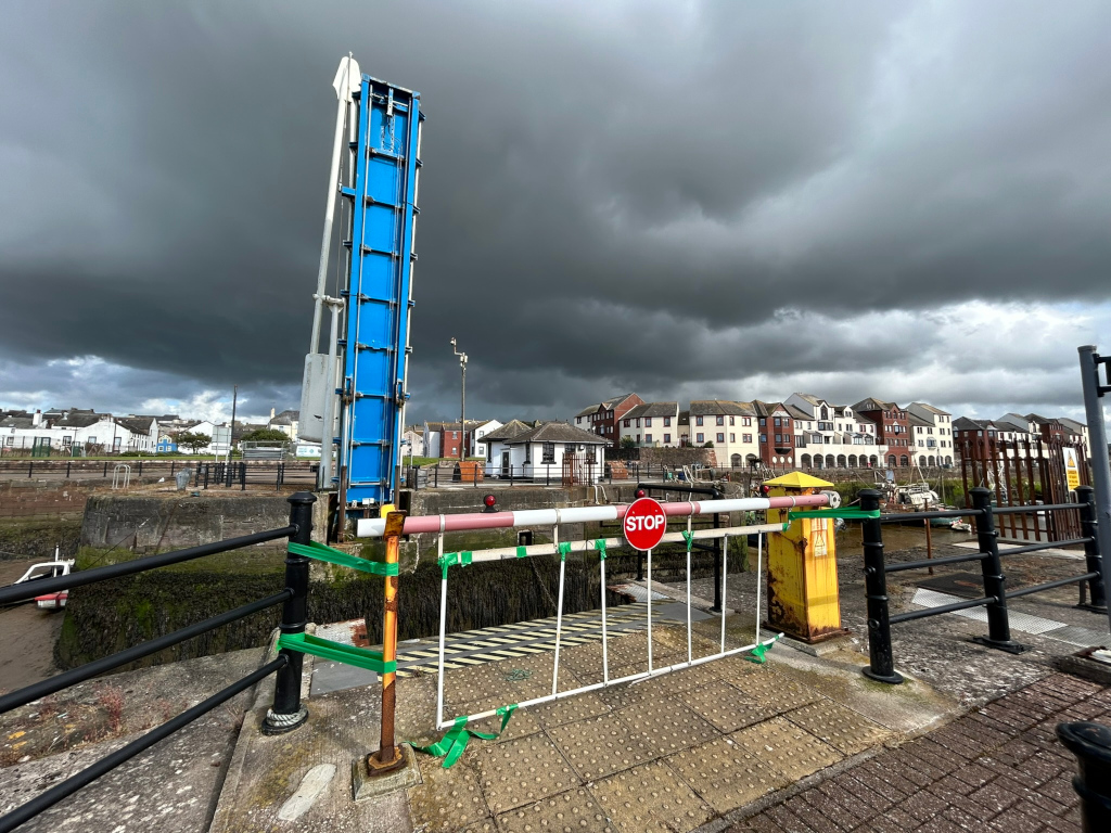 Closed gate at a harbour or marina, under a dramatic, dark sky filled with storm clouds. A tall blue hydraulic lift gate is prominent in the foreground, with a STOP sign on the barrier. The background features a row of multi-story buildings lining the waterfront. The overall mood is one of impending storm or recent bad weather.