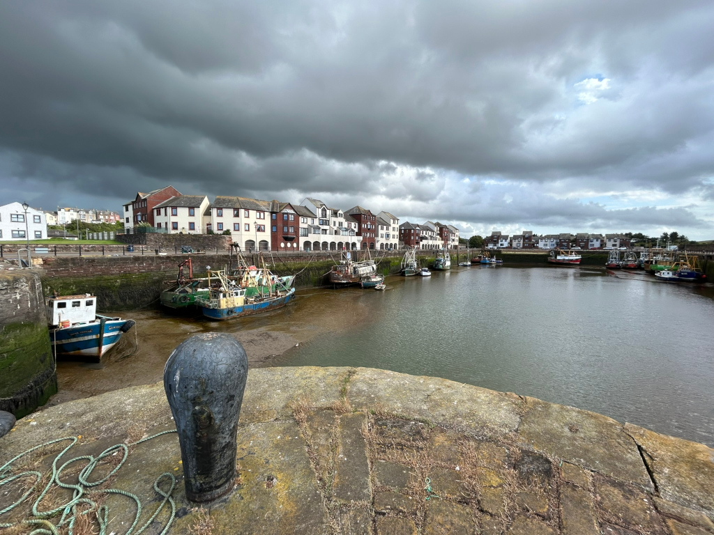 Harbor scene under a dramatic, cloudy sky. Numerous fishing boats are moored in the harbor, some appearing older and weathered. A row of multi-story buildings lines the harbor's edge. The overall atmosphere is somewhat somber and moody due to the dark clouds. The foreground features a weathered stone wall with a bollard and rope.