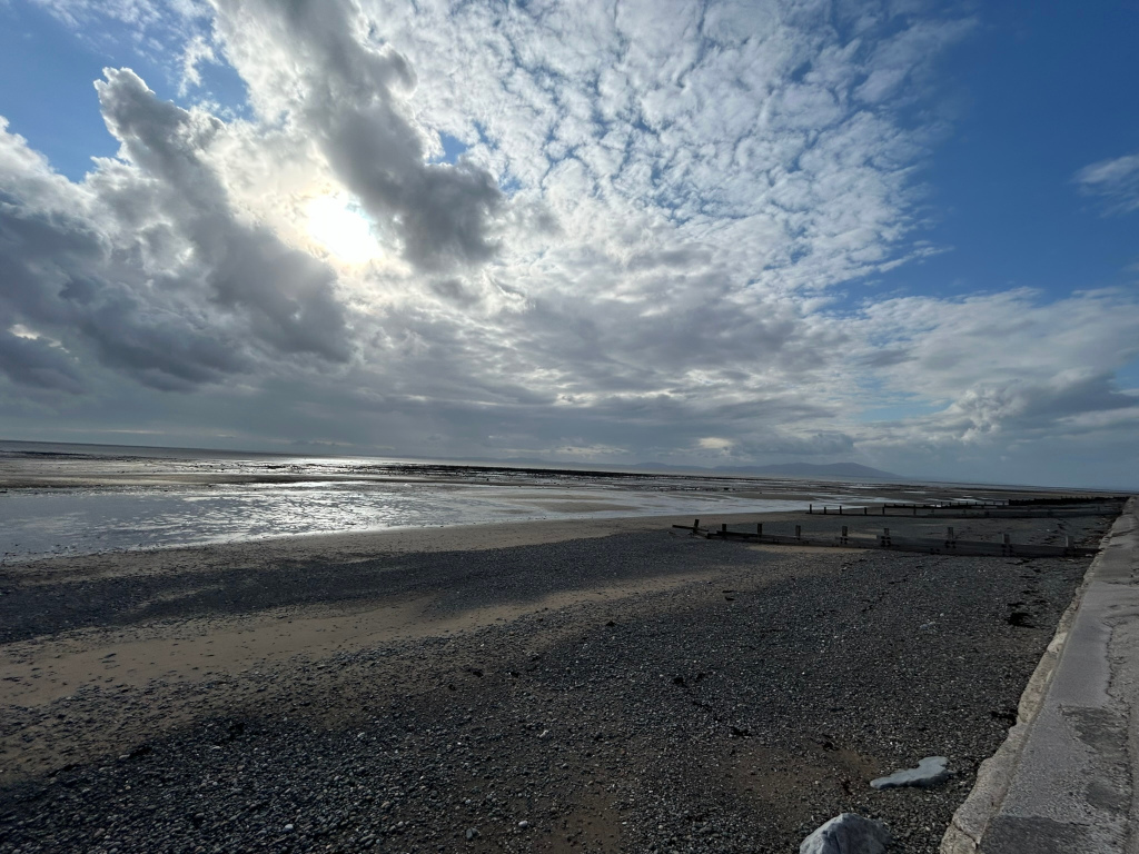 Beach scene under a dramatic sky. The sky is filled with a mix of fluffy white and darker gray clouds, with sunlight breaking through in places. The beach is composed of dark pebbles and sand, and a low-lying, weathered wooden structure stretches along the shoreline. A calm, reflective body of water lies between the beach and the distant horizon, where a faint landmass or mountain range is visible. The overall atmosphere is serene yet somewhat moody, reflecting the contrast between the bright sunlight and the darker cloud formations.