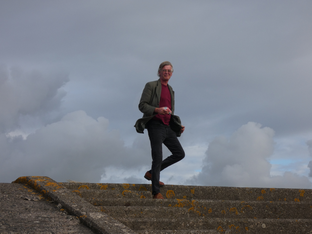 Charlie with glasses, wearing a maroon shirt and a grayish-green jacket, walking down a set of outdoor concrete stairs. The sky is overcast with large, fluffy clouds. Charlie appears relaxed and is carrying something small in his hand. The overall mood is calm and somewhat contemplative. The image's simplicity puts the focus on Charlie and his interaction with the space.