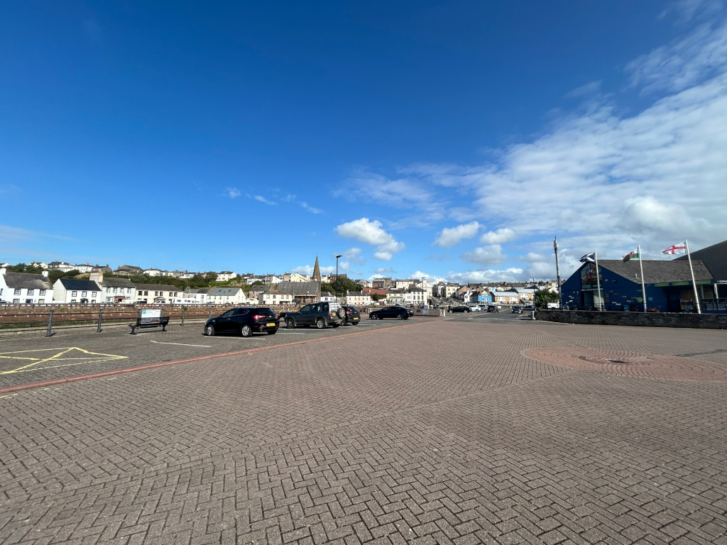 Mostly empty paved plaza or parking lot in front of a row of buildings. The buildings are multi-storied, mostly white or light-coloured, typical of a coastal town. A few cars are parked in the lot. In the background, slightly elevated, are more buildings in this same style. The sky is a bright, clear blue with some scattered white clouds. Several flags are visible on a building on the right. The overall impression is a sunny day in a small, likely seaside, town.
