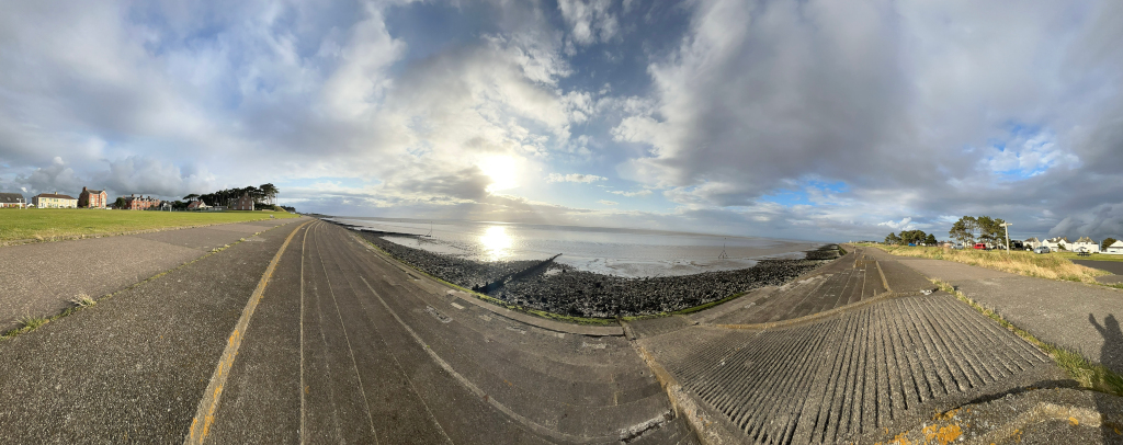 Panoramic view of a coastal scene. The foreground shows a paved walkway leading down to a rocky shoreline. In the background, there's a calm body of water under a partly cloudy sky. Buildings and some greenery are visible along the coast on either side of the walkway. The overall feel is serene and peaceful.