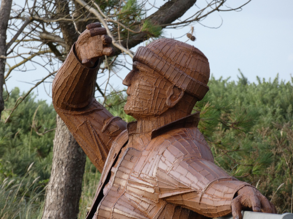 Close-up of a rusty-brown metal sculpture depicting a person, possibly a man, wearing a knitted hat and jacket. The figure is shown from the waist up, with one arm raised, as if shading their eyes or looking into the distance. The texture of the sculpture is rough and the style is somewhat abstract, with the clothing and facial features suggested rather than realistically depicted. The background is blurred but shows a natural setting with trees and shrubs.