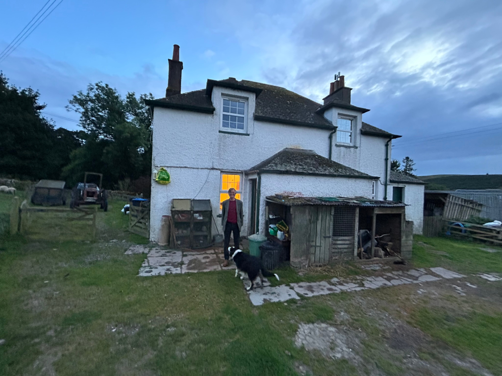 White farmhouse at dusk or twilight. Charlie stands in the doorway, with a border collie at his feet. Farm equipment and animal enclosures are visible in the yard. The overall scene is tranquil and evokes a sense of rural life.