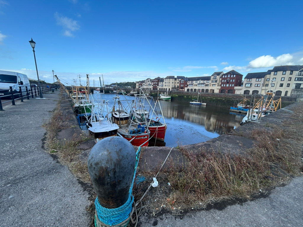 Harbor scene, likely in a coastal town. Several fishing boats are moored along a quay, their colourful hulls contrasting with the muted tones of the stone buildings lining the harbor. The water is relatively calm, reflecting the buildings and sky. A paved walkway runs alongside the quay, and a lamppost is visible in the background. 