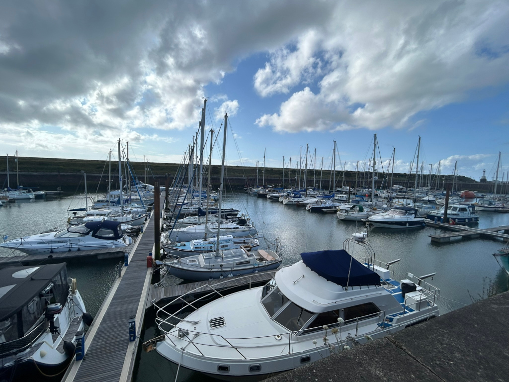 Marina filled with numerous sailboats and motorboats of varying sizes. The boats are moored along docks, with a wooden walkway visible between some of them. The sky is partly cloudy, with blue sky showing through the clouds. The overall scene is peaceful and suggests a calm day at a marina.