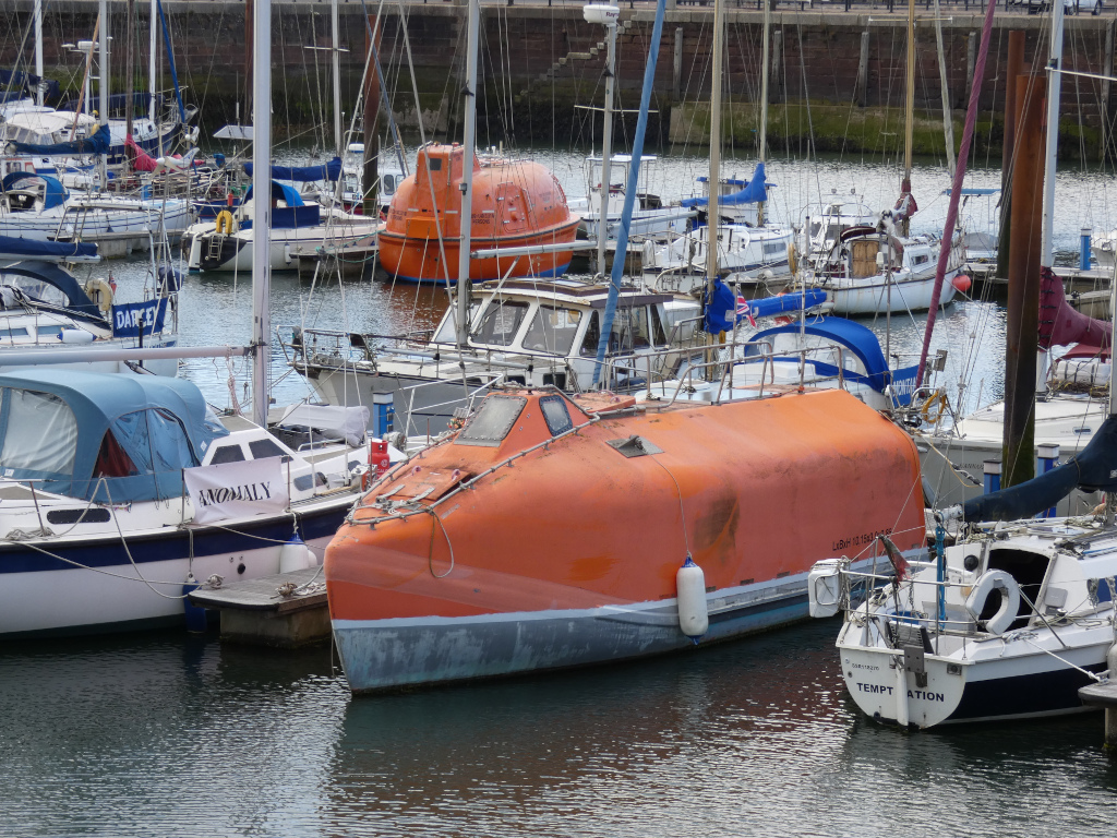 Marina filled with various sailboats and motorboats. Most prominent is a large, bright orange lifeboat, seemingly repurposed as a small boat. It is situated among other vessels, some with visible names like Anomaly and Temptation. The overall setting suggests a calm day in a harbour.