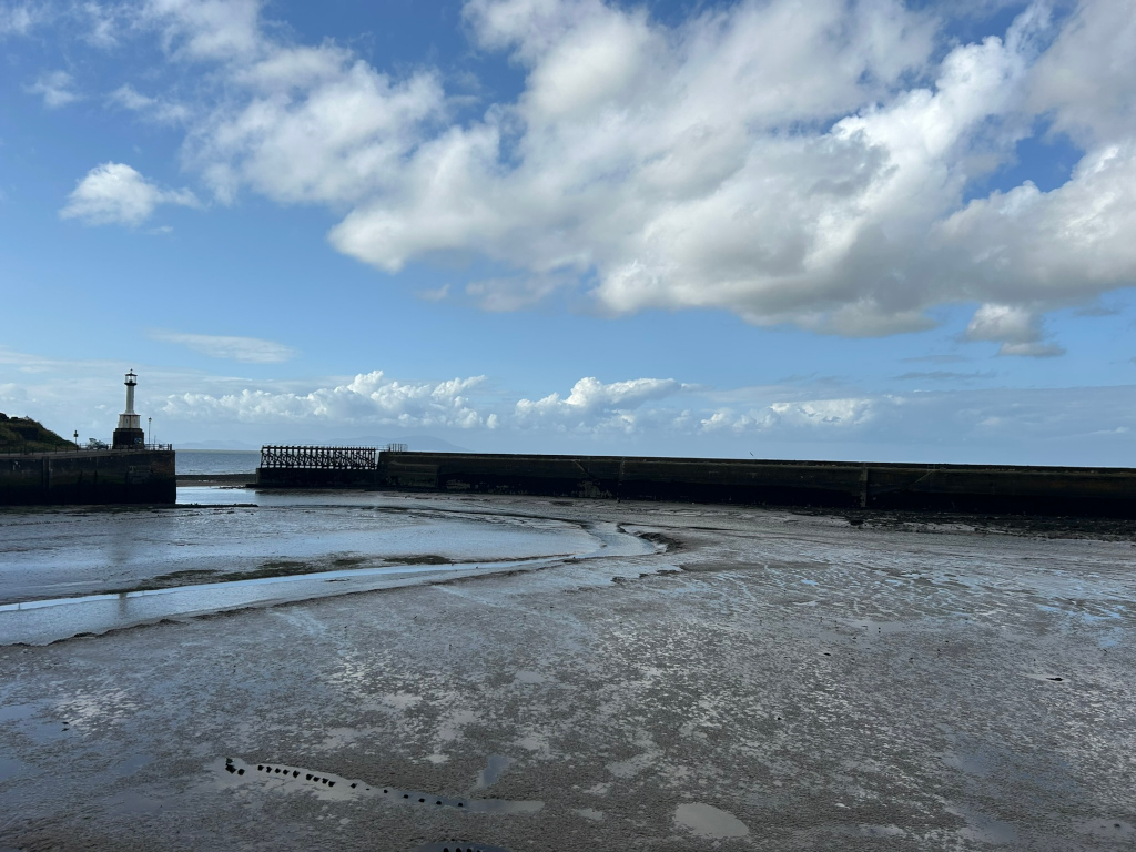 Low-tide scene at a harbour. A small lighthouse stands on one side of the harbour entrance, which is partially closed by a low, dark wall and a wooden structure. The foreground is expansive, muddy tidal flats reflecting the sky. The background features a calm sea line under a partly cloudy blue sky.