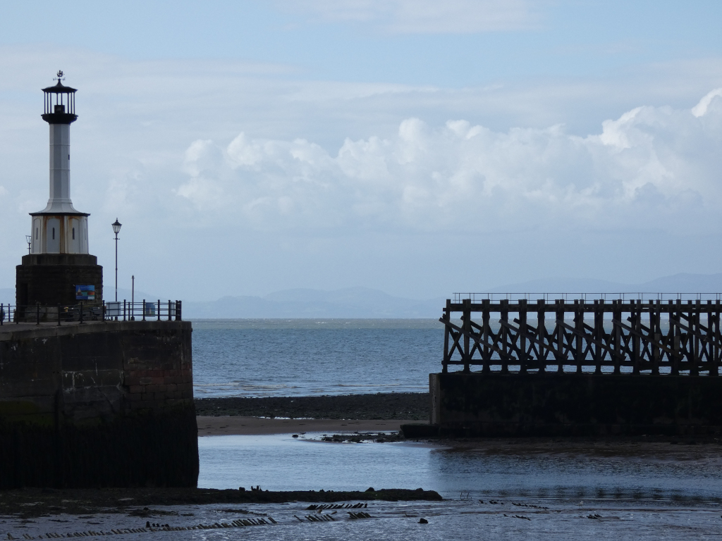Coastal scene featuring a small lighthouse standing on a stone structure to the left, with a section of an old wooden pier extending from the right into the calm sea. The background shows a tranquil sea and a distant shoreline under a partly cloudy sky. The overall mood is serene and slightly melancholic, hinting at a quiet, possibly historic, coastal location.