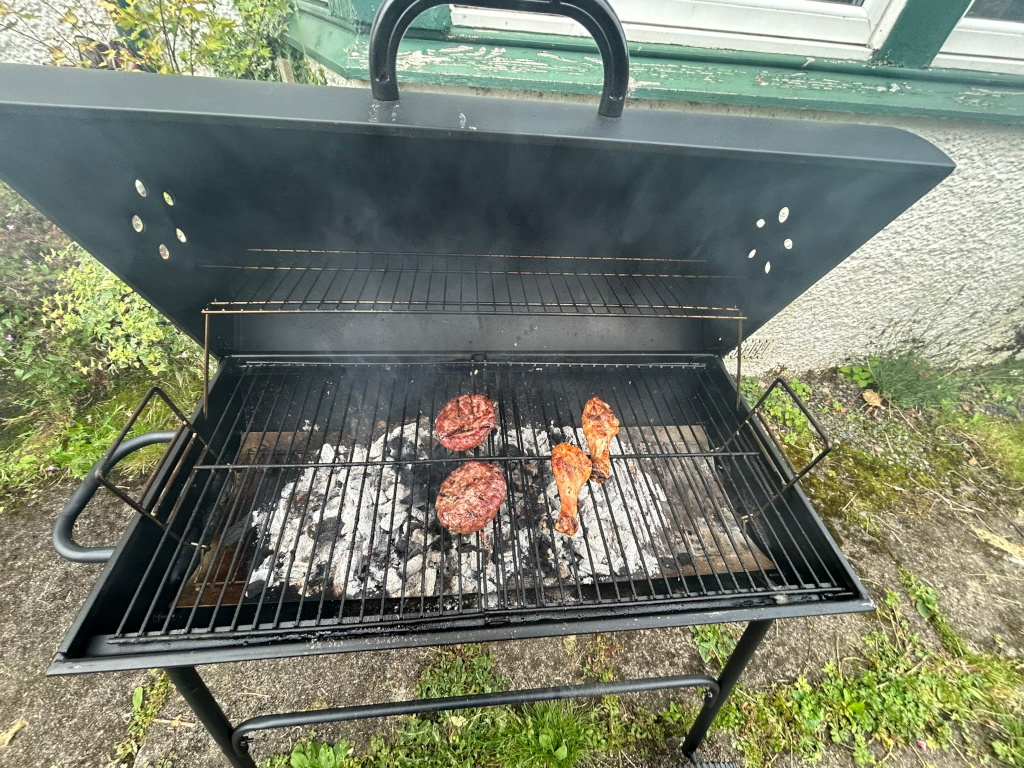 Black charcoal grill with two hamburger patties and two chicken drumsticks cooking on the grill. The grill is open, and some smoke is visible. The grill is outdoors on a paved surface with some grass and plants visible in the background.