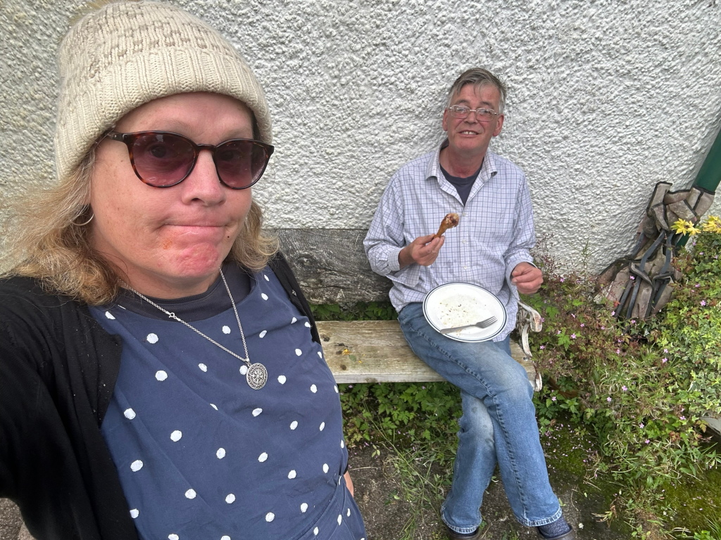 Selfie-style photograph of a woman and a man (Charlie) enjoying a meal outdoors. The woman, Leonie, is wearing a knit hat and sunglasses, is in the foreground. She's dressed in a navy blue polka-dot dress and cardigan. The man, wearing glasses and a plaid shirt, is sitting on a wooden bench behind her, eating what appears to be a piece of roasted chicken from a small plate. They are sitting against a whitewashed wall, with some greenery and plants visible in the background. The overall impression is one of casual outdoor dining.