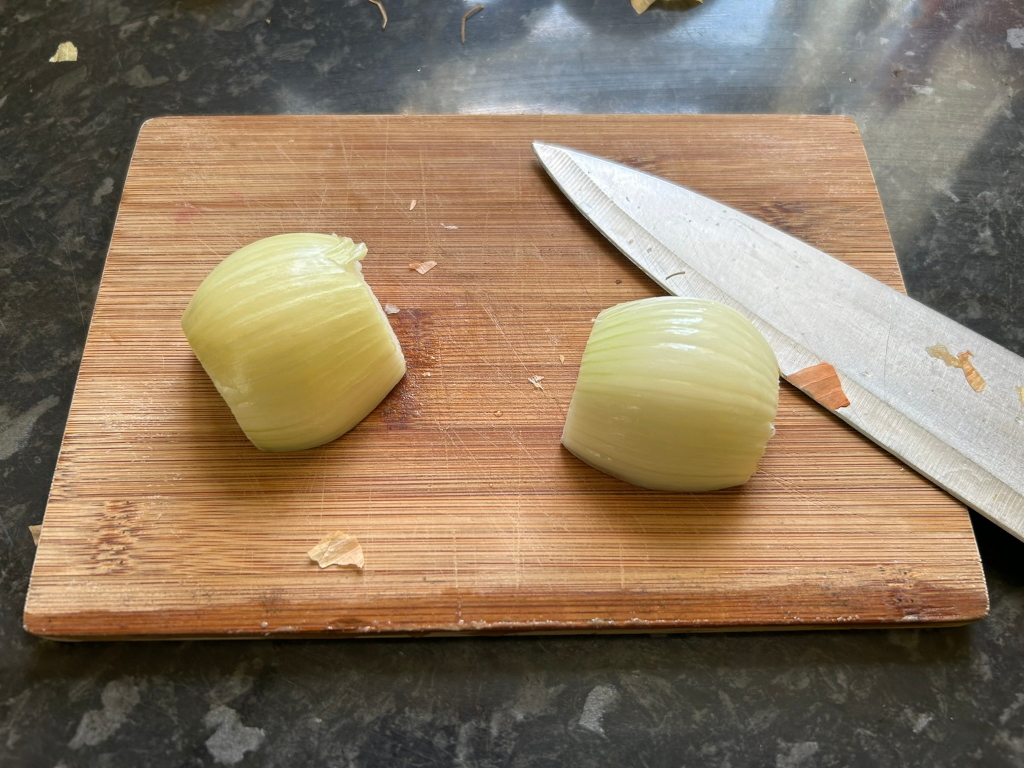 Bamboo cutting board with two quartered onions on it and a knife lying next to them. The onions appear to be freshly cut and are pale yellow in colour. The cutting board shows signs of previous use with some small bits of onion peel. The scene is simple and suggests food preparation is underway.