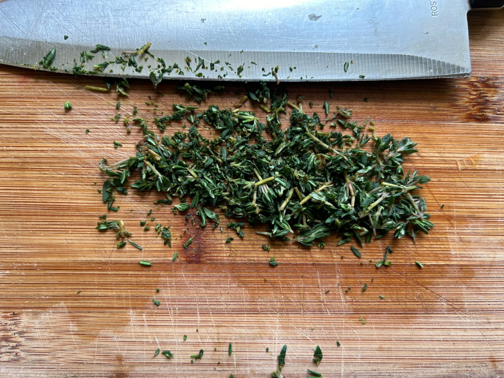 Pile of freshly chopped thyme leaves on a wooden cutting board, with a kitchen knife resting above it. The thyme is finely chopped, and some small pieces are scattered around the main pile. The cutting board shows signs of previous use.