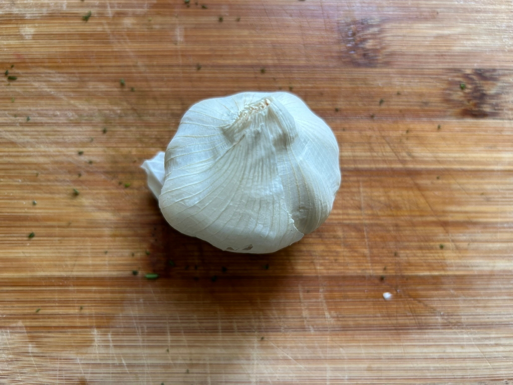 Single bulb of garlic, its papery outer skin mostly removed, sitting on a light brown wooden cutting board. The board shows signs of previous use, with visible scratches and small bits of debris. The focus is sharply on the garlic bulb.