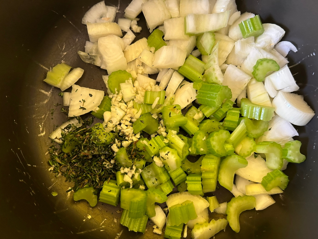 Dark-coloured pot containing chopped onions, celery, garlic, and thyme. The vegetables appear to be prepped for cooking, likely as part of a savoury dish.