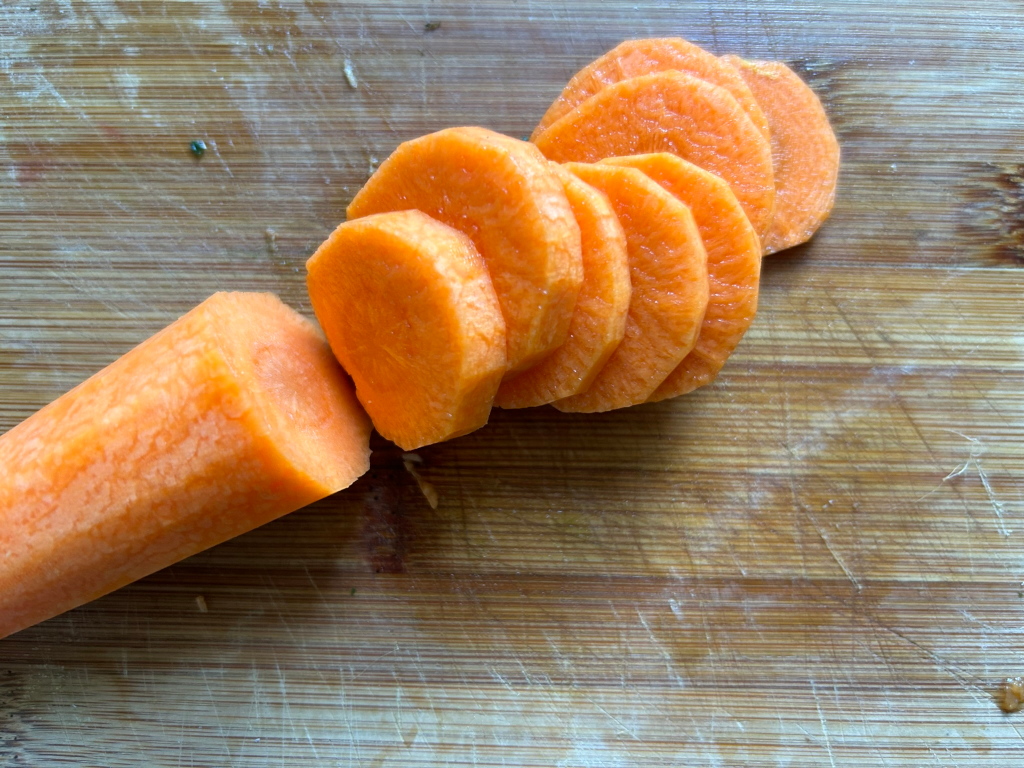 Carrot that has been peeled and sliced into several rounds. The carrot slices are arranged on a wooden cutting board. A larger piece of the carrot remains, next to the slices. The overall impression is simple and straightforward, focusing on the freshly cut carrot.