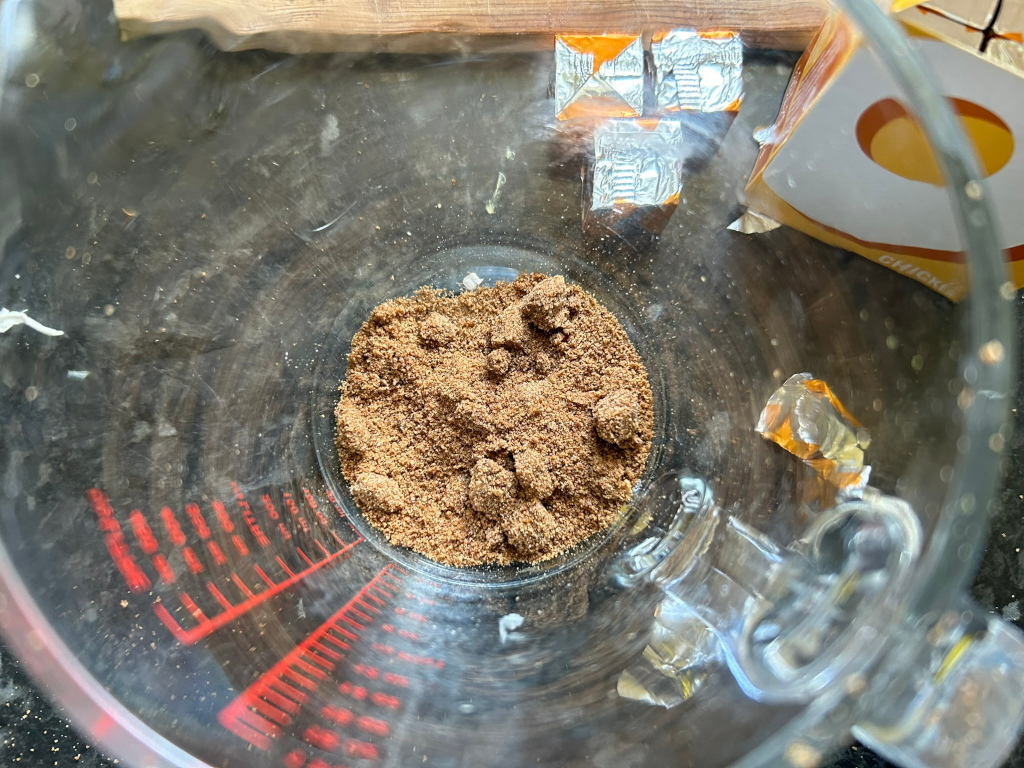 Glass measuring cup containing a brown, granular substance, possibly a spice blend or ground food. In the background, several foil packets of chicken bouillon cubes are visible, suggesting the brown substance might be related to a recipe using these cubes.