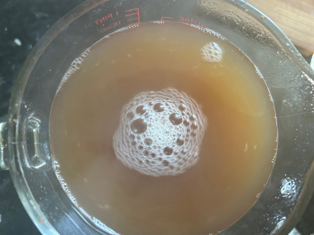 Glass measuring cup filled with a light brown liquid. A distinctive pattern of bubbles is visible in the centre of the liquid, seemingly created by a swirling or vortex motion. The liquid appears somewhat translucent. The cup shows measurement markings.