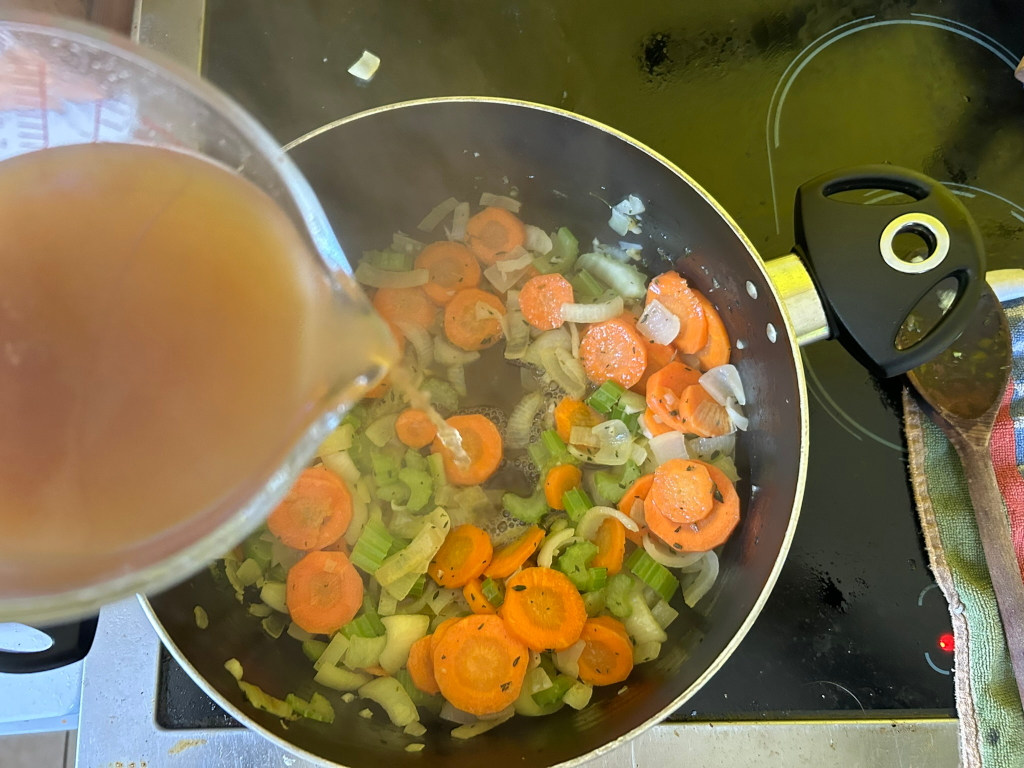 Leonie pouring broth into a pan containing sauteed carrots, celery, and onions. The pan is sitting on a stove top.