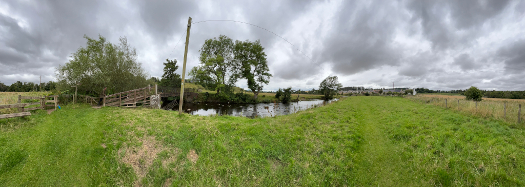Panoramic view of a tranquil rural scene. A small wooden footbridge spans a calm stream or river, flanked by lush green pastures. Trees provide shade along the banks and a cloudy sky dominates the background. In the distance, hints of more developed areas or houses are visible. The overall impression is one of peaceful countryside.