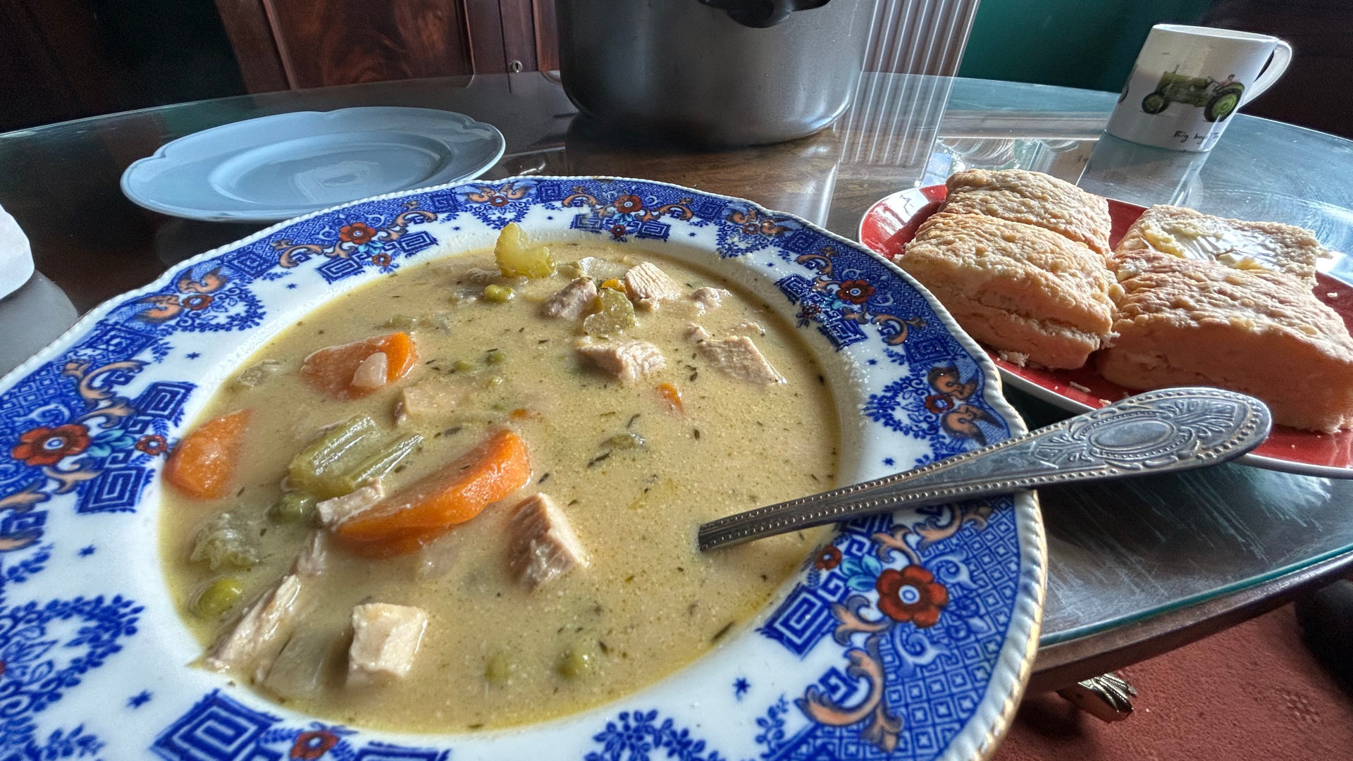 A bowl of creamy chicken soup, garnished with carrots and peas, served in a vintage blue and white patterned bowl. Next to it, on a red plate, are several pieces of what looks like buttery bread or biscuits. A silver spoon rests in the soup. In the background is a large, grey pot (likely the soup's source) and a mug. The setting appears to be a home dining table. The overall impression is one of a comforting, home-cooked meal.