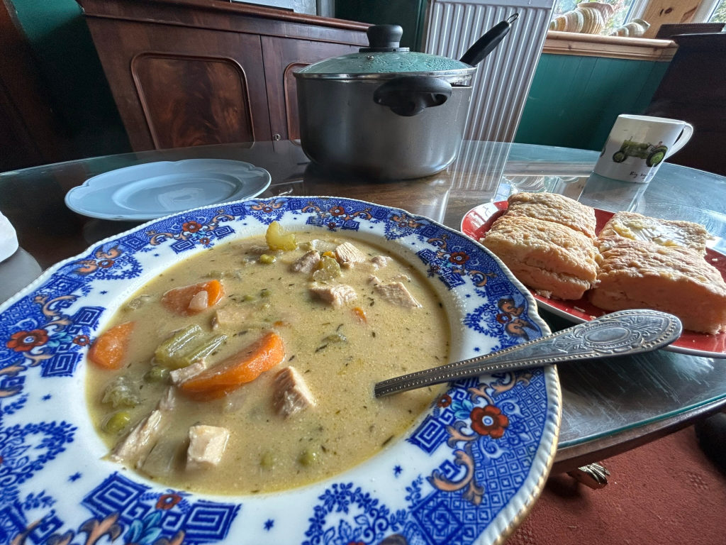 Bowl of creamy chicken soup, garnished with carrots and peas, served in a vintage blue and white patterned bowl. Next to it, on a red plate, are several pieces of what looks like buttery bread or biscuits. A silver spoon rests in the soup. In the background is a large, grey pot (likely the soup's source) and a mug. The setting appears to be a home dining table. The overall impression is one of a comforting, home-cooked meal.