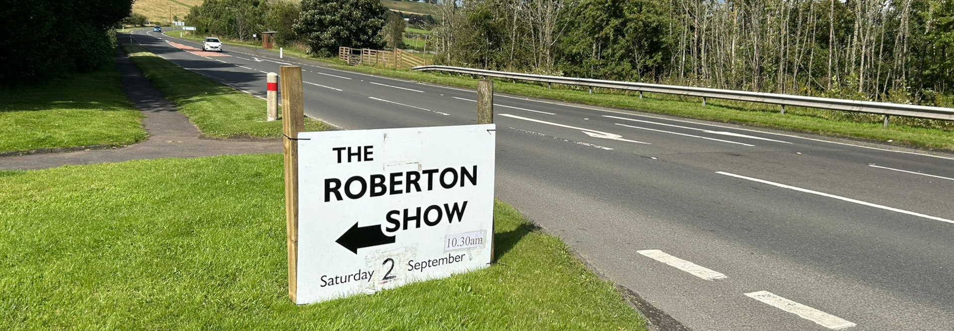 Roadside sign advertising The Roberton Show on Saturday, September 2nd, starting at 10:30 am. A left-pointing arrow indicates the direction to the show. The sign is situated beside a country road with green fields and trees visible in the background.