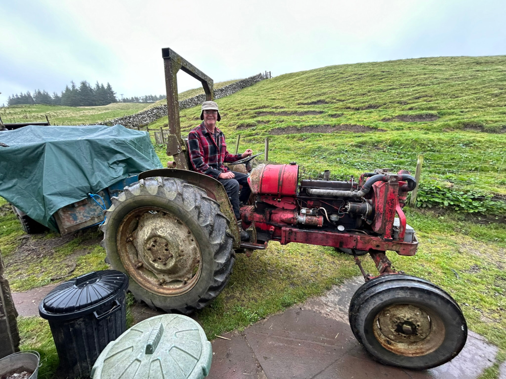 Charlie sitting on an old, red tractor. He appears to be a farmer, wearing a plaid shirt and a hat. The tractor is somewhat weathered and rusty, suggesting it's been used extensively. It's parked on a grassy area near a stone wall, with a covered trailer visible behind it. There are also some trash cans in the foreground. The overall setting appears to be a rural farmland.