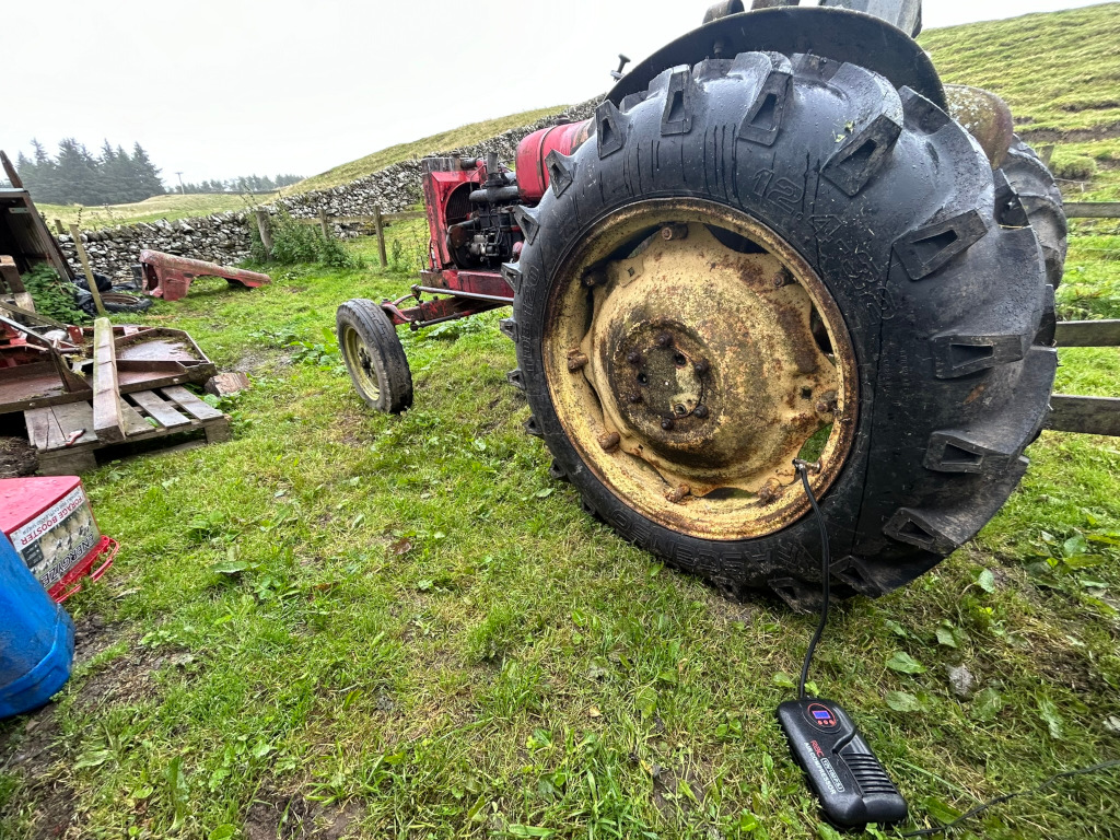 Red vintage tractor parked in a grassy field, next to a stone wall.  The focus is on the tractor's rear tire, which appears worn and is connected to a portable tire inflator. Surrounding the tractor are various pieces of farm equipment and debris. The overall mood is one of rustic work and possibly repair. The background is a somewhat overcast or misty landscape.