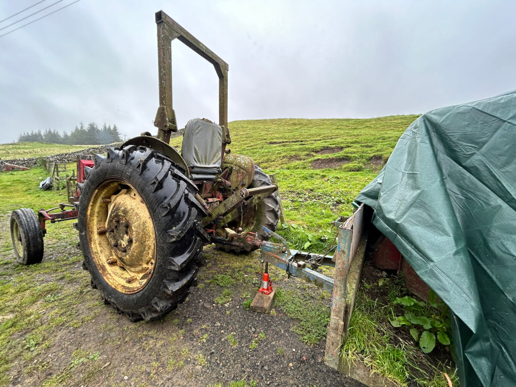 Old, rusty tractor parked in a field, attached to a piece of farm equipment covered by a tarp. The setting appears rural and somewhat overcast. The tractor shows signs of age and wear, suggesting it may be vintage or no longer in regular use. 
