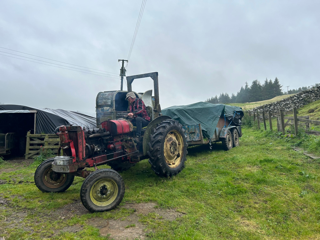 Charlie  sitting on an old, rusty red tractor pulling a trailer covered by a tarp.  The tractor and trailer are parked on a grassy field. A stone wall and some trees are visible in the background, suggesting a rural setting. The sky is overcast. The overall impression is one of rural life and possibly farm work.