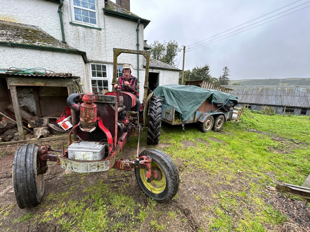 Charlie with short greying hair wearing a plaid shirt and a hat, sitting on a vintage red tractor. The tractor is parked in front of a rustic white farmhouse. The tractor is attached to a small, tarp-covered trailer. The setting appears to be rural, with overgrown grass and other farm-related items visible in the background. The overall mood is serene and evokes a sense of rural life and possibly a feeling of nostalgia associated with older farming equipment.