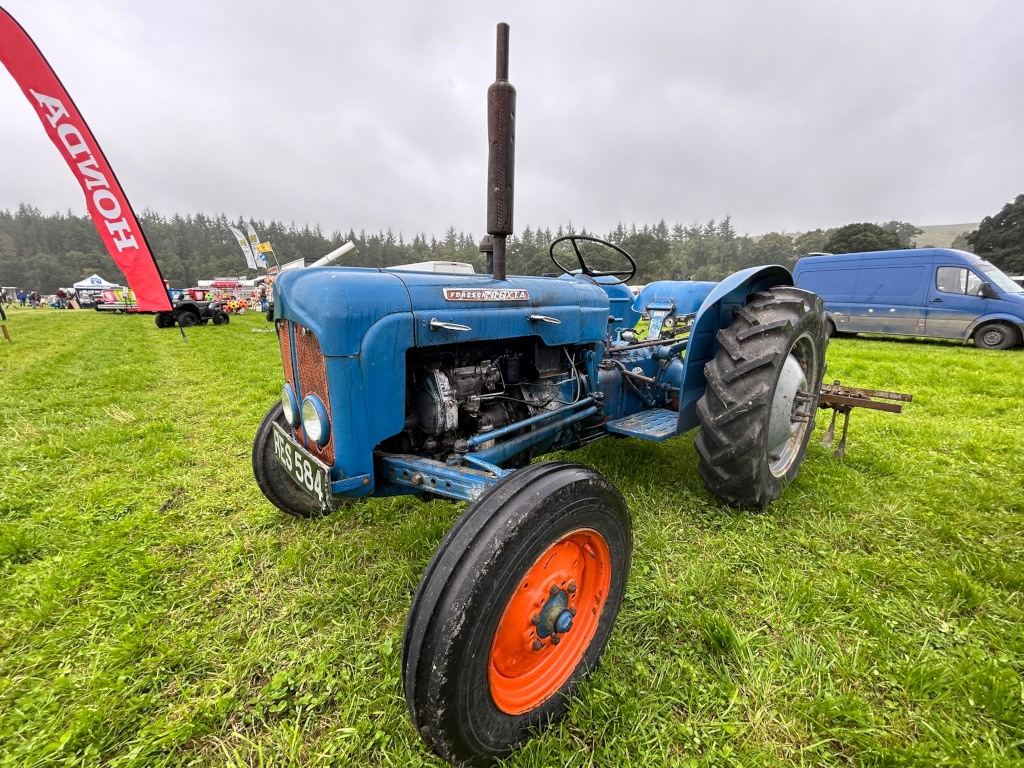 Vintage blue Fordson Dexta tractor parked on a grassy field. The tractor appears to be well-maintained, and its paint job is relatively intact. The background includes other vehicles, suggesting a rural setting, possibly a fair or agricultural show. A Honda banner is visible in the far left background.