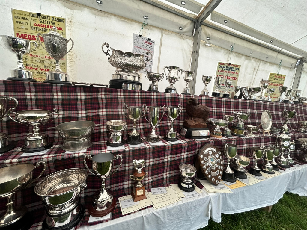 Large collection of silver trophies and awards displayed on a table covered with a red and black plaid tablecloth. The trophies vary in size and style, suggesting a range of competitions and achievements. Several vintage posters advertising past events, possibly agricultural shows, are visible behind and amongst the awards, adding historical context. The scene suggests a display at a rural fair or agricultural show, celebrating past winners.