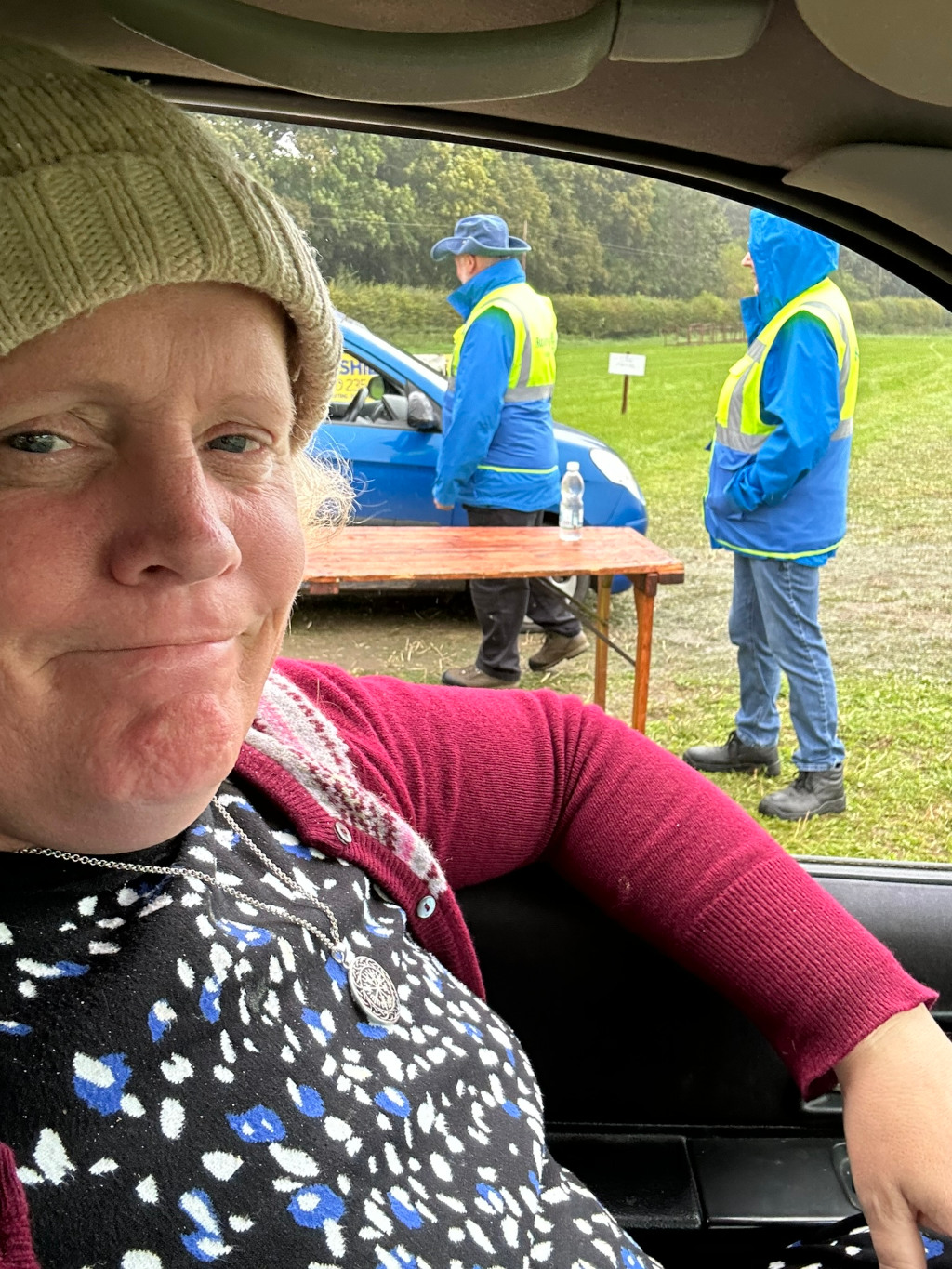 Selfie taken from inside a car. The person taking the photo is a woman wearing a beige knitted hat and a burgundy cardigan over a black and blue patterned top. She's wearing a silver necklace with a circular pendant. In the background, out of focus, are two people wearing bright blue jackets and high-visibility vests, standing by a small wooden table next to a blue car in a grassy field. The weather appears overcast. The overall impression is of a casual outdoor event, possibly a volunteer activity or community gathering. The woman in the foreground seems relaxed and perhaps a bit amused, contrasting with the more formal appearance of the people in the background.