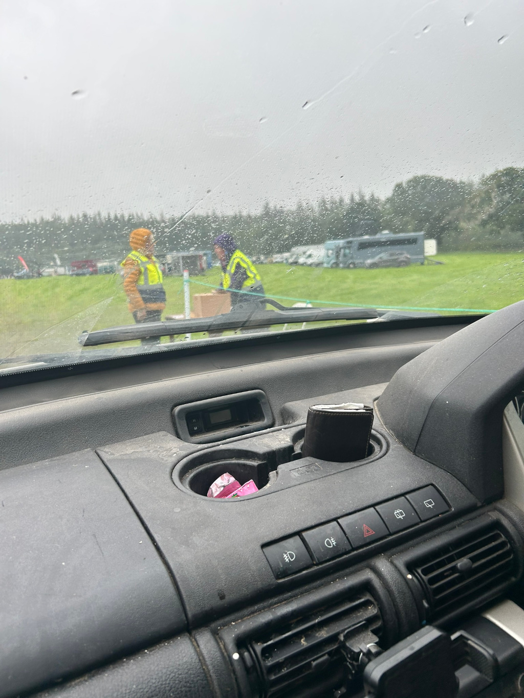 View from inside a car, looking out at a rainy scene. Two people wearing high-visibility vests are visible in the mid-ground, seemingly engaged in some activity near a box. The background shows a grassy field with parked vehicles and a line of trees under a grey, overcast sky. The car's interior is dusty, and a wallet rests in the cup holder. The overall impression is one of a somewhat bleak, possibly outdoor event taking place in inclement weather.