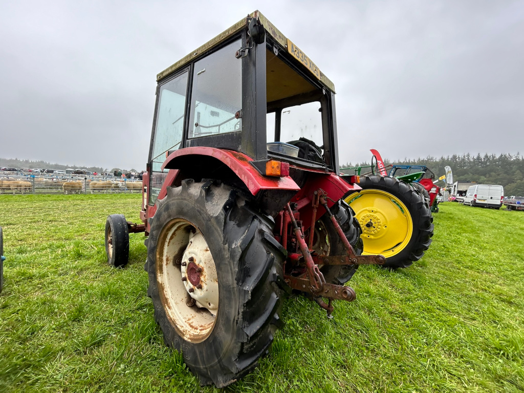 Rear view of a vintage red tractor, predominantly taking up the frame, parked on a grassy field. The tractor appears to be at an agricultural show or similar event, with sheep pens visible in the background and other farm machinery partially in view.