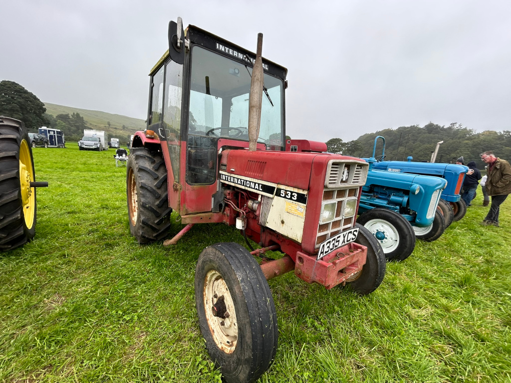 Red International 533 tractor prominently in the foreground. Behind it are two blue tractors, slightly out of focus, suggesting a display or gathering of vintage farm machinery. The setting appears to be a grassy field, possibly at a rural event, with a few other vehicles and people visible in the background. The overall atmosphere is calm and pastoral.