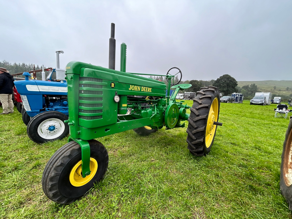 Vintage John Deere tractor, predominantly green, prominently displayed in a grassy field. Another, older, blue tractor is visible in the background, along with several other vehicles and people in the distance, suggesting a rural setting possibly at a show or gathering. The overall impression is one of nostalgia and agricultural heritage.