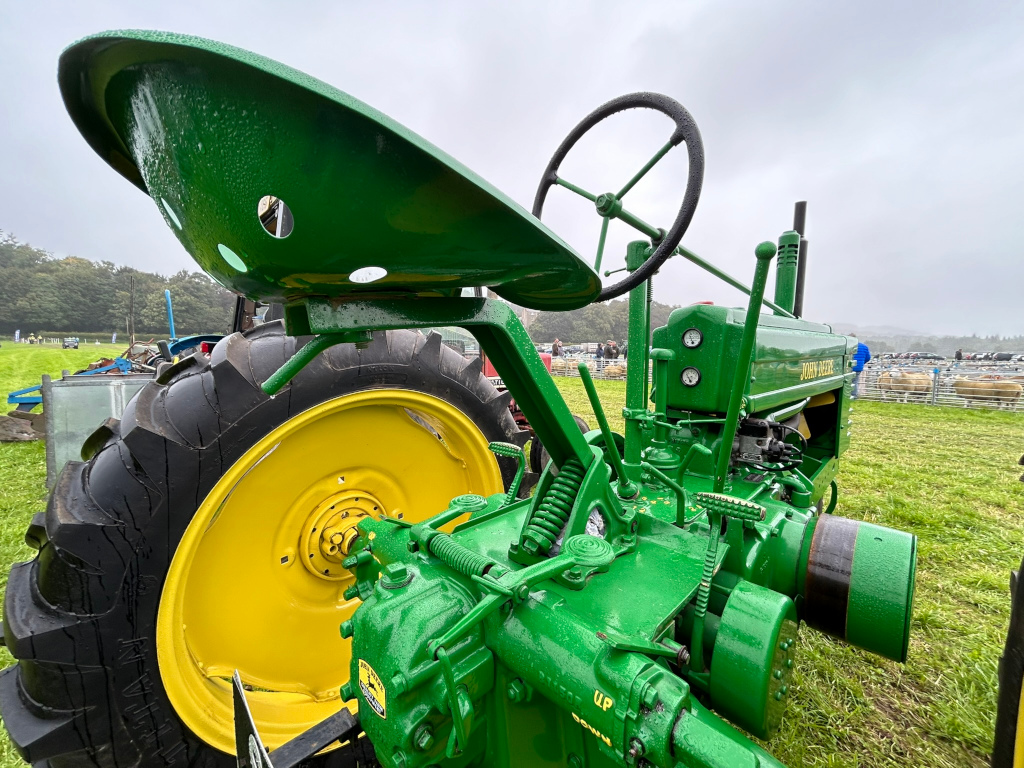 Close-up view of a vintage John Deere tractor, specifically the rear and side, partially covered in raindrops. The tractor is green and yellow, with visible mechanical details and a large rear tire. The setting appears to be an outdoor agricultural event or show, with sheep pens visible in the background under a cloudy sky.