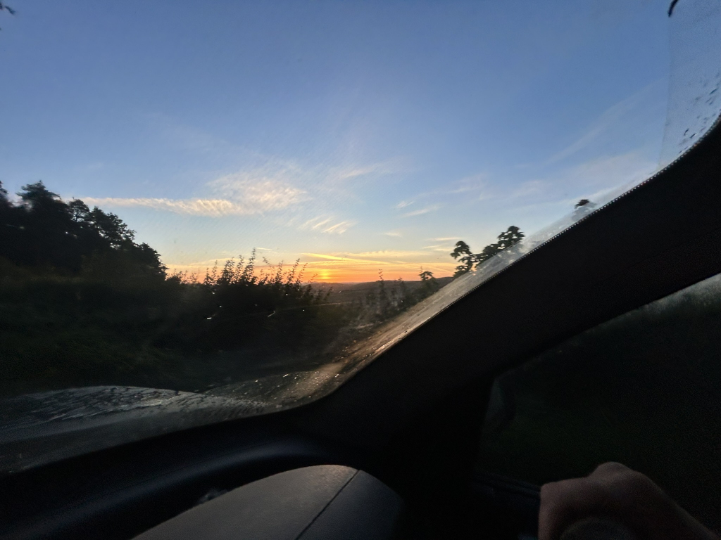 Sunrise viewed from inside a car. The sun is rising behind a line of trees, and the sky is a mix of blue, orange, and pink. The car's windscreen is dirty, adding a slightly hazy effect to the view. A hand is visible in the bottom right corner, suggesting someone is driving.